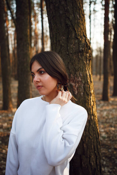 a young beautiful girl in a white sweater stands near a tree and looks into the distance in the fores