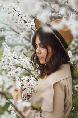 Bej ceketli ve şapkalı bir kadın parkta ya da çiçek açan bir elma ağacının yanında duruyor.
