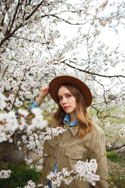 Parkta poz veren bej ceketli ve şapkalı bir kız ya da çiçek açan bir elma ya da kayısı ağacının yanında duran bir kız.