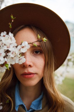 Parkta poz veren bej ceketli ve şapkalı bir kız ya da çiçek açan bir elma ya da kayısı ağacının yanında duran bir kız.