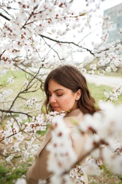 Parkta poz veren bej ceketli bir kadın ya da çiçek açan bir elma ya da kayısı ağacının yanında duran bir kadın.