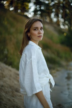 Beyaz keten elbiseli genç kız ormanın bulanık arka planına bakıyor. Şefkat ve gençlik