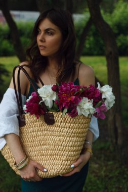Parkın ortasında duran esmer bir kadının çantasındaki beyaz ve kırmızı şakayık demeti.