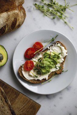 Krem peynirli domatesli tam tahıllı ekmeğin avokado tostu ve mikroyeşil. Beyaz mermer bir yüzey üzerinde bir tabakta sandviç. Vejetaryen kahvaltı