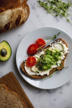 Krem peynirli domatesli tam tahıllı ekmeğin avokado tostu ve mikroyeşil. Beyaz mermer bir yüzey üzerinde bir tabakta sandviç. Vejetaryen kahvaltı