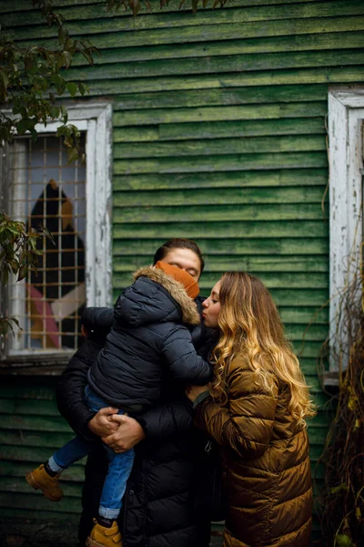 dad is holding the baby in his arms and mom is trying to kiss him outdoors. a young family in warm clothes stands near an old green wooden house. family walk in cold weather
