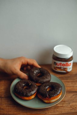 KYIV, UKRAINE - 1 Nisan 2021: çikolatalı çörek ve bir kutu Nutella. Elin çikolatalı çörek tutuyor. Nutella ile donuta uzan..