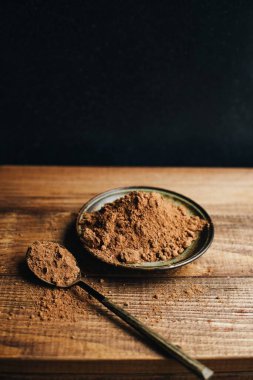 Küçük bir tabakta kakao tozu. Kakao tozu ve kaşık. Ahşap bir masada pişirme malzemesi. Siyah arka planda kakao tozu.
