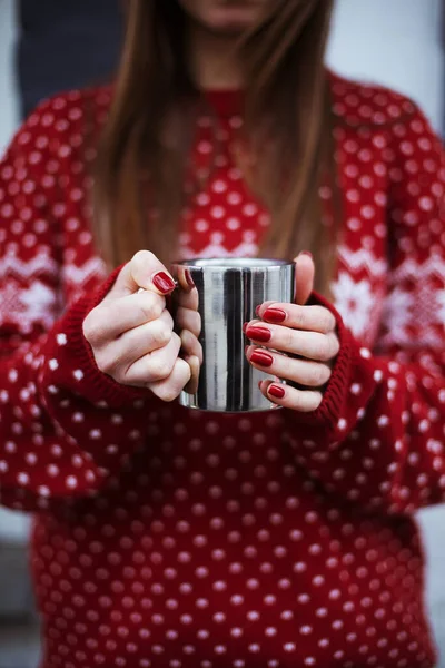 a steel mug in the hands of the girl. woman in red Christmas sweater. hot tea or coffee or cocoa drink or mulled wine in a steel cup. the girl warms her hands with a hot mug.