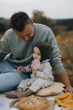 Baba ve bebek dışarıda. Babam parkta battaniyenin üzerinde oturan bebeğine hayran. Genç baba çocuğa bakıyor. Ev yapımı turtayla sonbahar pikniği