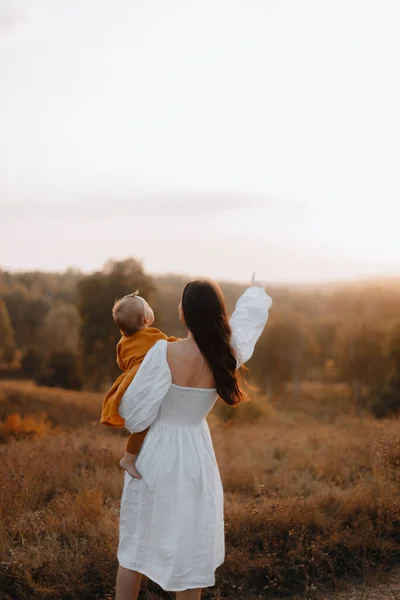 mother with baby in her arms points into the distance. a young mother holds a little girl in a brown dress in her arms. mother and daughter stand with their backs in the field at sunset. young woman