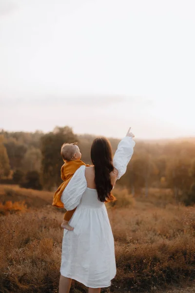 抱着孩子的母亲走到远方 一位年轻的母亲抱着一个穿着棕色衣服的小女孩 日落时 母女俩背着背站在田里 女青年 — 图库照片