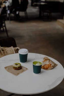 İki bardak sıcak içecek lütfen. Paket matcha latte. Kafedeki beyaz bir masada içkili iki kağıt bardak. Badem kruvasan ve çörek..