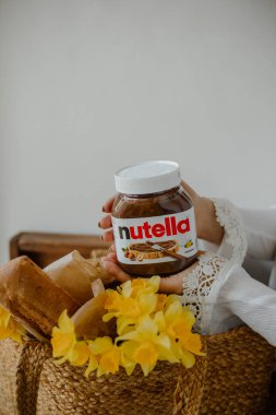 KYIV, UKRAINE - 1 Mayıs 2021: Kadın ellerinde fındık kavanozu. Kız çantasına bir kutu Nutella koyuyor. Nergisli çanta ve beyaz arka planda ekmek..
