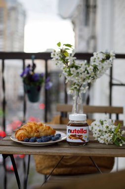 KYIV, UKRAINE - 1 Mayıs 2021: Balkon masasında Nutella kavanozu. Ay çöreği ve yaban mersini, çiçek açan dalları olan bir vazonun yanında bir tabakta. Balkonda yemek ve çiçeklerle süslenmiş bir masa..