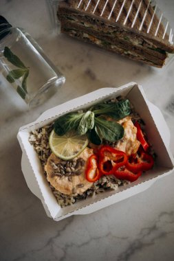 Kırmızı balık etli beyaz ve kahverengi pirinç, doğranmış kırmızı biber, tohum ve bir dilim limon. Beyaz mermer bir masada beyaz kağıt bir kabın içinde yemek. Kilo vermek için sağlıklı yiyecek teslimatı.