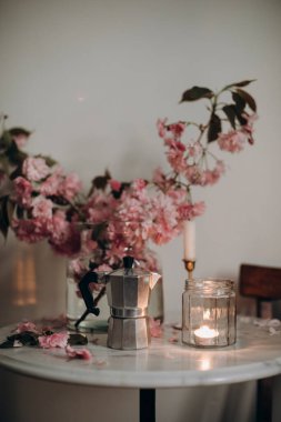 Çelik İtalyan Moka Tenceresi mermer kahve masasında pembe çiçeklerle. Şeffaf bir kavanozda yanan mum mermer bir masada, sakura dallarıyla dolu bir vazoda..