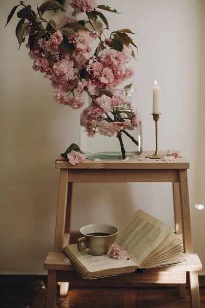 Şeffaf vazosu ve pembe dalları olan küçük tahta bir merdiven. Şeffaf bir vazoda sakura dalları ve bir fincan kahveyle açık bir kitap. Pembe sakura yaprakları. Boşluğu kopyala