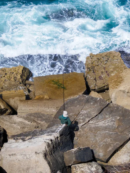 Alone fisherman with hat fishing in the sea from a cliff. Waves, ocean, fishing pole