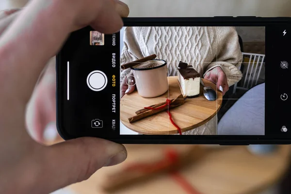 Akıllı telefon ekranında yemek fotoğrafı var. Elleri akıllı telefon tutuyor ve tahtanın üzerinde kahve ve kek fotoğrafları çekiyor..