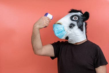 Lateks inek maskesi takan kişi pembe arka planda kızıl ötesi termometreyle ateşini ölçüyor.