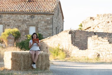 Kırsal kesimde orta yaşlı esmer Latin bir kadın, arka planda taştan bir evin olduğu saman balyasında oturan turuncu kapaklı bir tablete danışıyor..