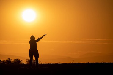 silhouette of a woman on a bale of straw in a field with her arms outstretched as if she were offering something with a large sun and mountains in the background