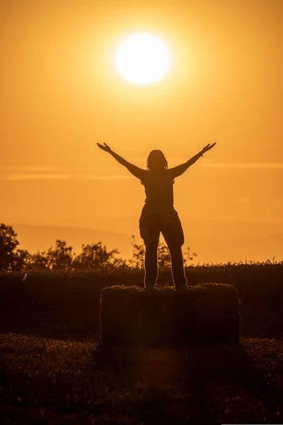 backlit photograph of the silhouette of a woman perched on a bale of straw with her arms outstretched and the sun behind her during the golden hour. Rural scene.