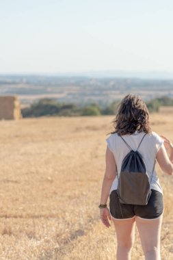 Orta yaşlı esmer kadın şort ve beyaz tişört giyiyor, sırtında siyah bir sırt çantası olan hasat edilmiş bir tarlada yürüyor. Arka planda saman balyaları yığını.