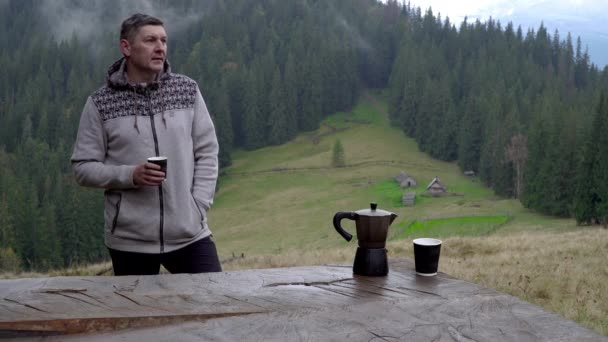Мужчина пьет кофе и смотрит на красивый пейзаж. Кофеварка и кружка на столе — стоковое видео