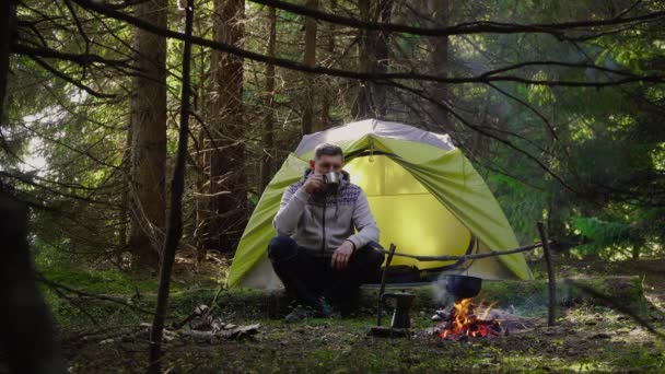 A man drinks coffee near a tent in the forest — Stock Video