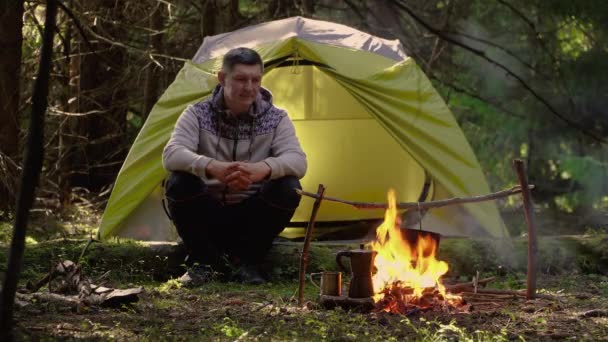 Een man drinkt koffie bij een tent in het bos. — Stockvideo