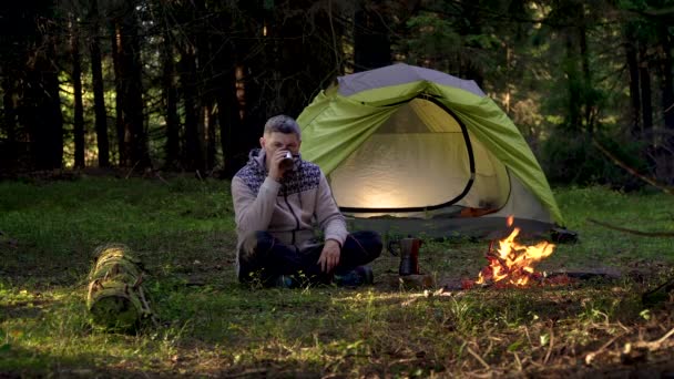 Ένας άντρας πίνει καφέ κοντά σε μια σκηνή στο δάσος. — Αρχείο Βίντεο