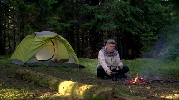 Мужчина пьет кофе возле палатки в лесу — стоковое видео