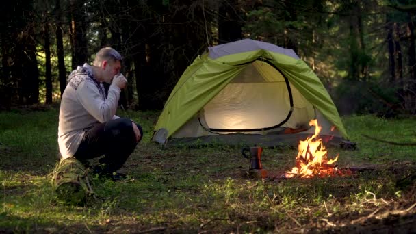 En man dricker kaffe nära ett tält i skogen — Stockvideo