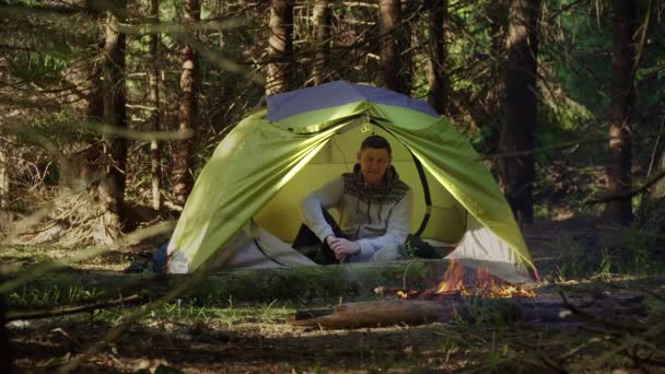 Een man in een tent bij een vuur in het bos — Stockvideo