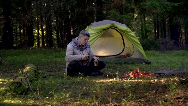 Ein Mann trinkt Kaffee in der Nähe eines Zeltes im Wald — Stockvideo