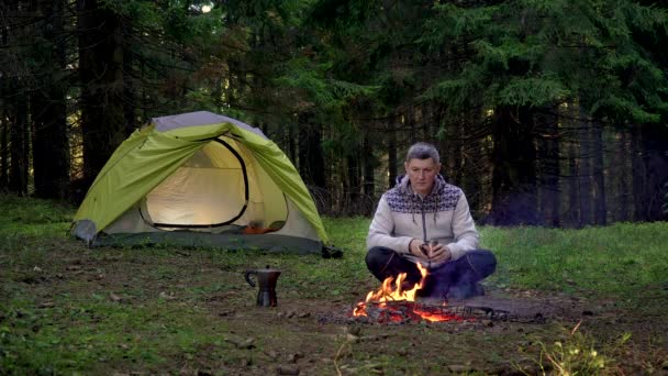 Um homem bebe café perto de uma tenda na floresta — Vídeo de Stock