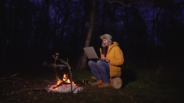 A young man works at a laptop near the fire in the forest — Stock Video