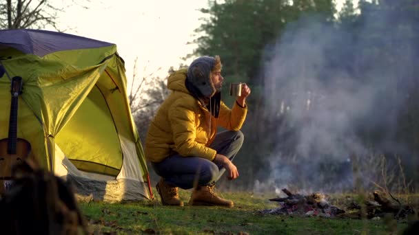 A man drinks tea near a campfire and a tent in the woods — Stock Video