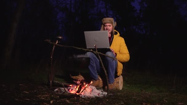 Молодий чоловік працює біля ноутбука біля багаття в лісі — стокове відео