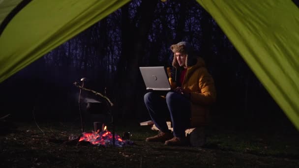 Ein Mann arbeitet am Laptop am Lagerfeuer im Wald — Stockvideo