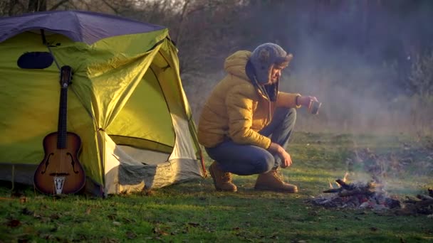 A man drinks tea near a campfire and a tent in the woods — Stock Video