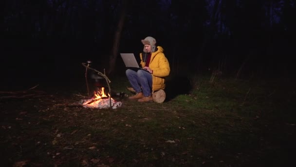 A young man works at a laptop near the fire in the forest — Stock Video