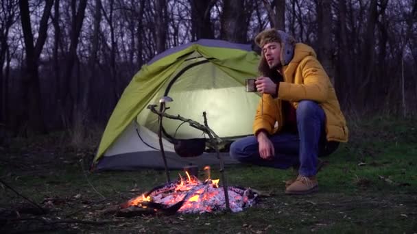 Мужчина пьет чай у костра и палатку в лесу — стоковое видео