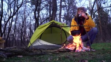 Bir adam kamp ateşi yanında çay içer ve ormanda çadır kurar.