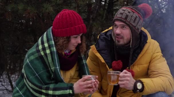 Junger Mann und Frau trinken Tee am Feuer im Winterwald — Stockvideo