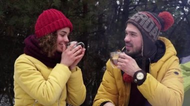 Genç adam ve kadın kış ormanında ateşin yanında çay içiyorlar.