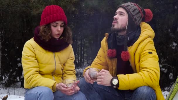 Молодой человек и женщина пьют чай у костра в зимнем лесу — стоковое видео