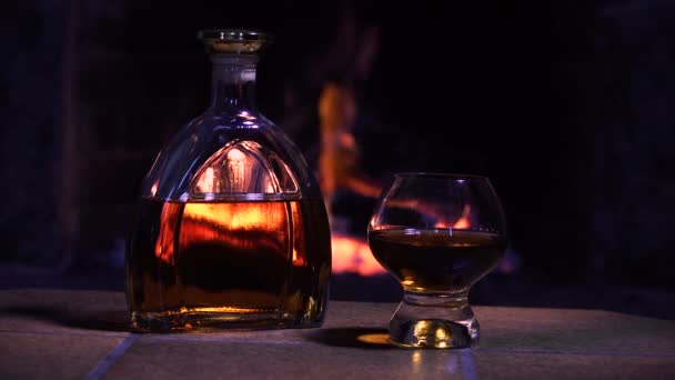Μπουκάλι και ποτήρι με ουίσκι ή κονιάκ στο φόντο της φωτιάς στο τζάκι — Αρχείο Βίντεο